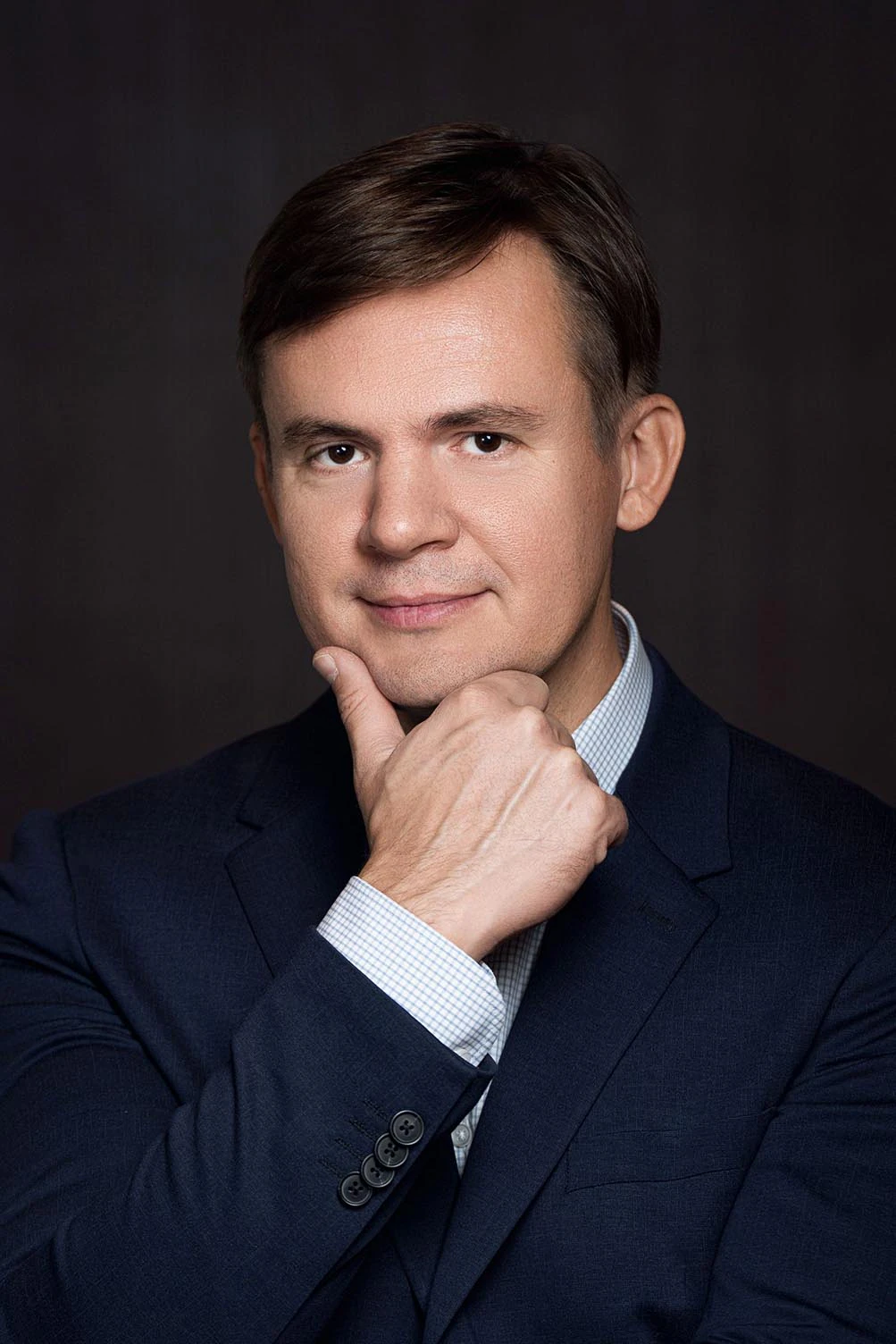 Вдумчивый деловой портрет: мужчина в синем костюме держит руку у подбородка, студийная съемка крупным планом на темном фоне.
